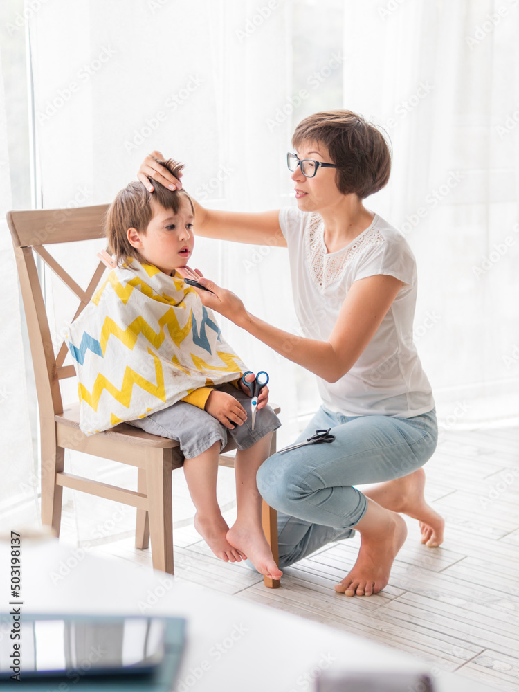 Mother cuts her son's hair by herself. Little boy sits, covered with cloth, and holds pair of scissors. Family life at home. Mother cuts her son's hair by herself. Little boy sits, covered with cloth, and holds pair of scissors. Family life at home.