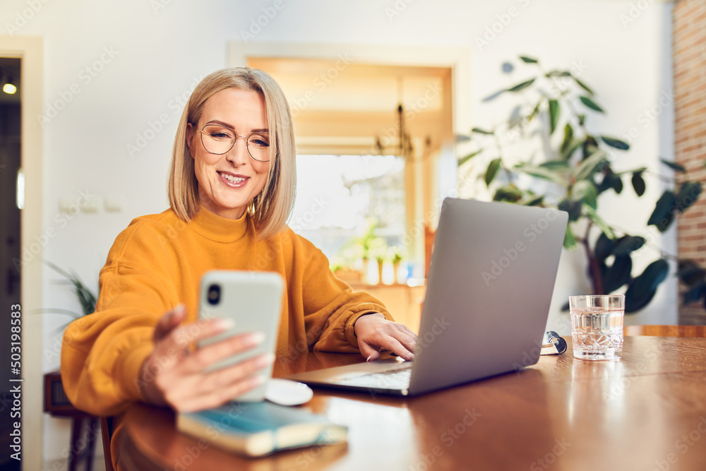 Smiling mature woman using smartphone working at home office with laptop sitting at dinning table Smiling mature woman using smartphone working at home office with laptop sitting at dinning table