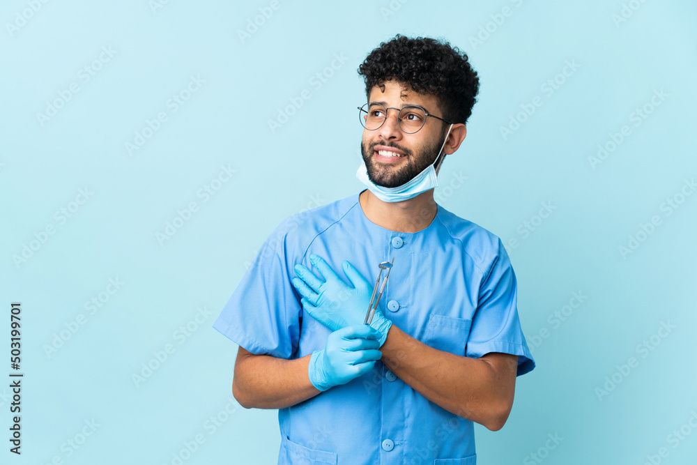 © luismolinero - Moroccan dentist man holding tools isolated on blue background looking up while smiling