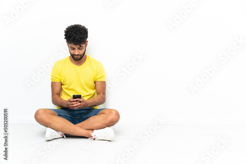 Young Moroccan handsome man sitting on the floor over isolated background sending a message with the mobile