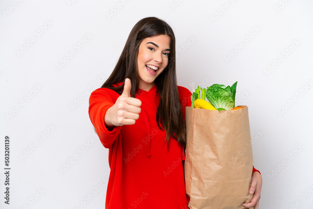 Young Brazilian woman holding a grocery shopping bag isolated on white background with thumbs up because something good has happened Young Brazilian woman holding a grocery shopping bag isolated on white background with thumbs up because something good has happened