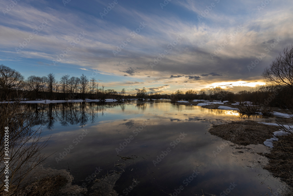 Epic spring landscape by the river. The flood of the river in early spring. A colorful sunset is reflected in the water. March evening landscape.