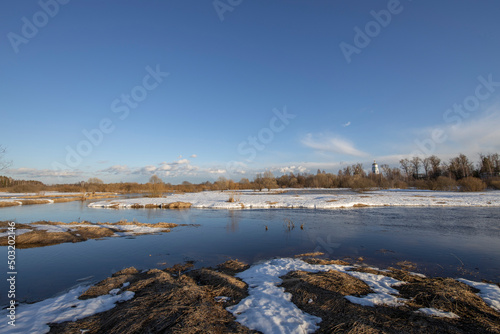 A picturesque landscape, early spring, a river with snow-covered banks, dry grass and bushes. March sunny day by the river. The first thaws, the snow is melting.