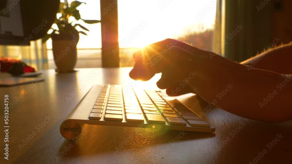 TRZIN, SLOVENIA, 20.5.2022: SLOW MOTION, CLOSE UP: Unrecognizable person working late on computer at sunset. Late afternoon sun shining over fingers of a person typing on wireless keyboard in office