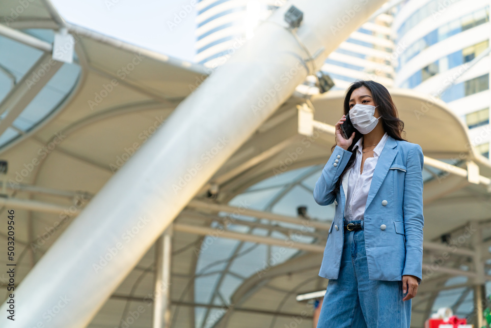 Obraz premium Asian businesswoman wearing face mask talking on cellphone walking city street in morning rush hour. Business woman working busy using mobile phone for working corporate business in office district.