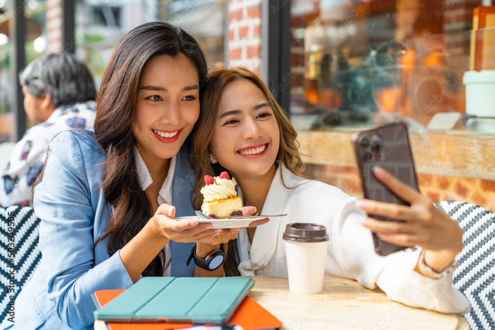 Asian woman friends using smartphone selfie together while sitting at outdoor coffee shop eating bakery and drinking coffee in urban city street. Beautiful female enjoy outdoor lifestyle in the city