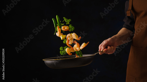 Fototapeta Naklejka Na Ścianę i Meble -  Shrimps with parsley and vegetables in a pan in a frozen flight on a black background. Asian cuisine, sea delicacies, gourmet food. Hotel, restaurant, cafe, picnic, banquet. Banner, advertising.