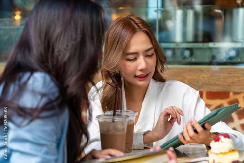  Two Asian businesswoman colleague discussion business plan while sitting at outdoor coffee shop urban city street. Happy business woman with using digital tablet working corporate teamwork together