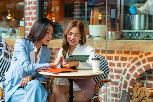  Two Asian businesswoman colleague discussion business plan while sitting at outdoor coffee shop urban city street. Happy business woman with using digital tablet working corporate teamwork together