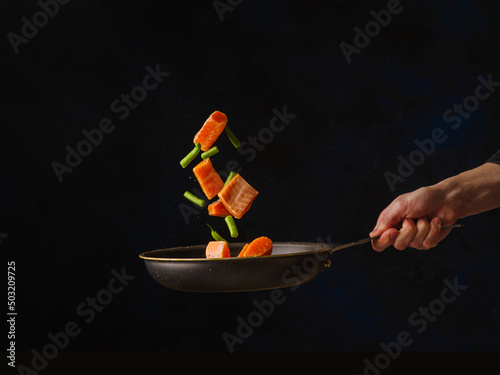 Pieces of red fish and vegetables on a frying pan in a frozen flight on a dark background. Minimalism. There is free space to insert. The process of cooking fish by a professional chef.