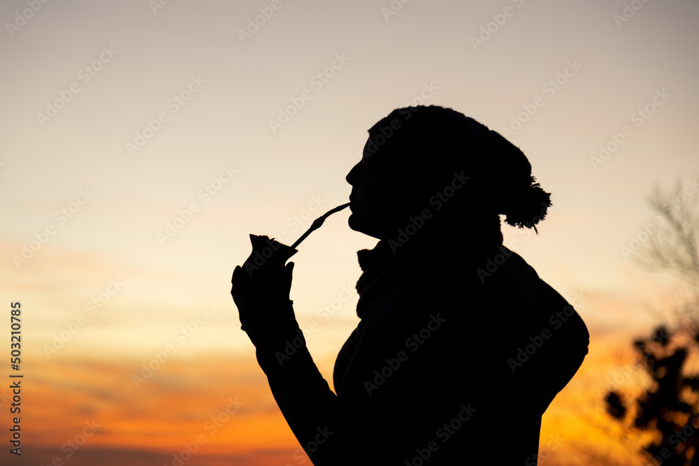 Silhouette of a woman with a cap drinking yerba mate, traditional South American drink, at sunset. Twilight sky