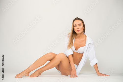 a young girl with a beautiful body in white underwear and a white shirt poses on a white background sitting in full growth