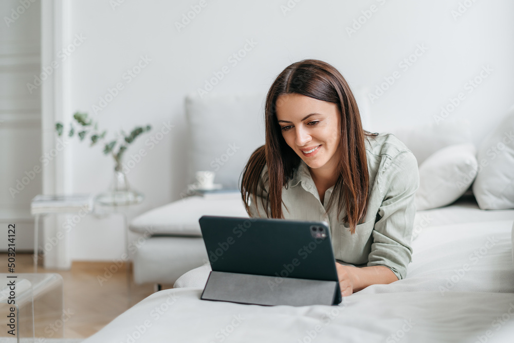 Happy pretty young woman in casual outfit sitting on sofa at home and using tablet while browsing internet