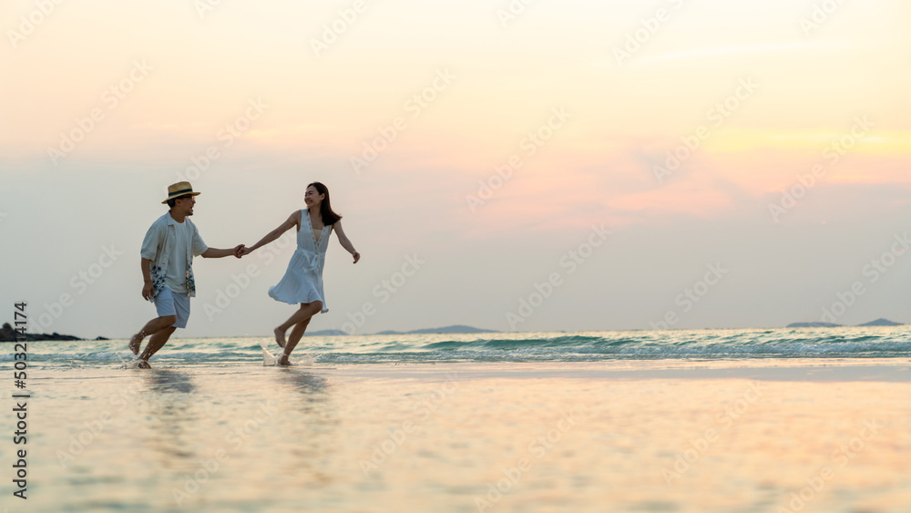 Fototapeta premium Happy Asian family couple walking and playing in sea water together on tropical beach at summer sunset on beach vacation. Husband and wife relax and enjoy outdoor lifestyle on holiday travel vacation