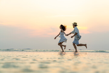 Happy Asian family couple walking and playing in sea water together on tropical beach at summer sunset on beach vacation. Husband and wife relax and enjoy outdoor lifestyle on holiday travel vacation