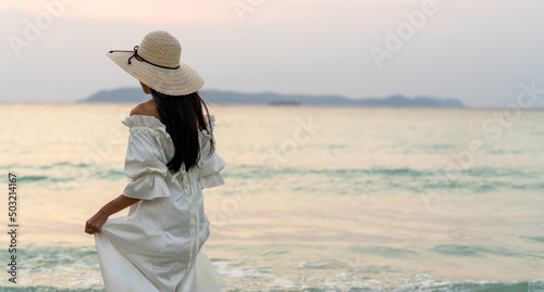 Young beautiful Asian woman in white dress walking on tropical island beach at summer sunset. Smiling female relax and enjoy outdoor lifestyle activity in beach holiday vacation and summer travel