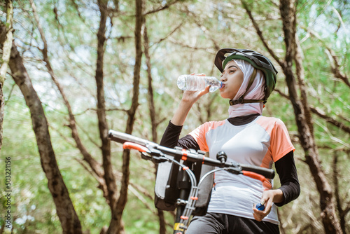 Woman in hijab drinking mineral water bottle when thirsty while cycling on outdoor background