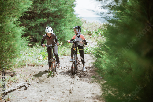 Two female cyclists pass through a sandy road on a beach with lots of trees