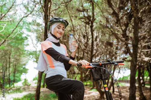 Attractive sporty woman in hijab holding bottle while cycling in sportswear and helmet on outdoor background