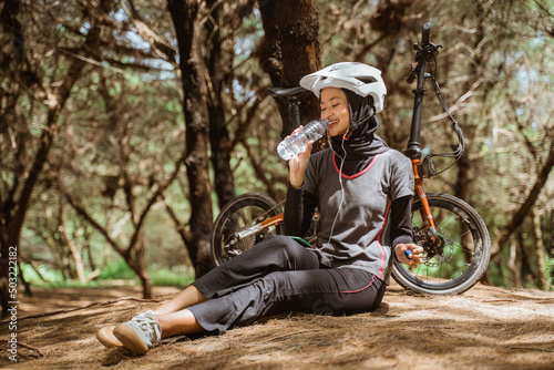 veiled girl wearing earphones while drinking with a bottle during a cycling break in the park