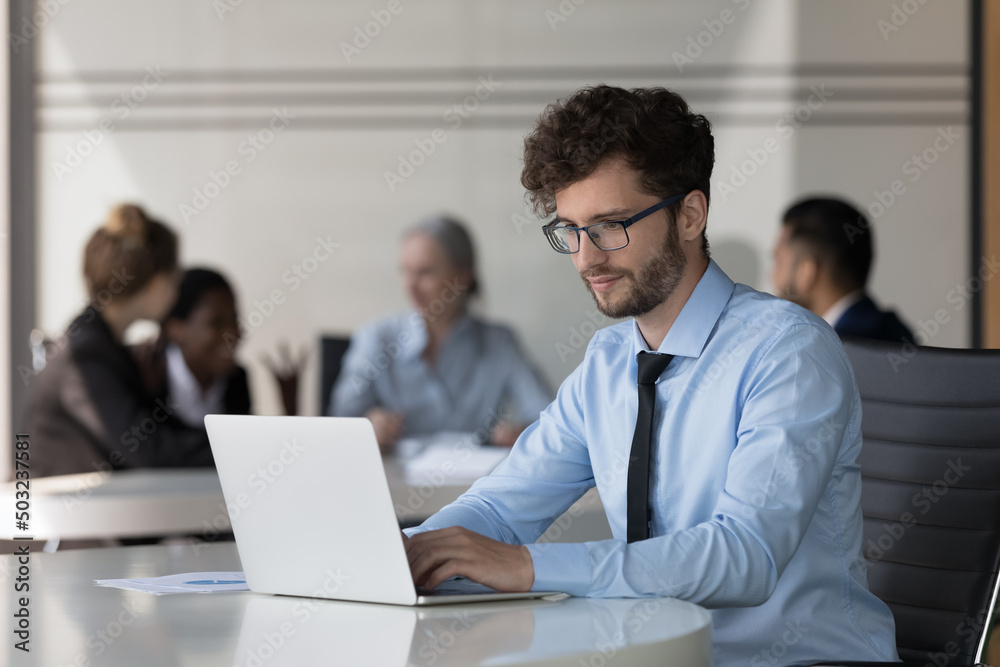 Serious young startup leader guy working on project in co-working space. Millennial businessman in glasses using laptop computer in office with corporate team in background Serious young startup leader guy working on project in co-working space. Millennial businessman in glasses using laptop computer in office with corporate team in background