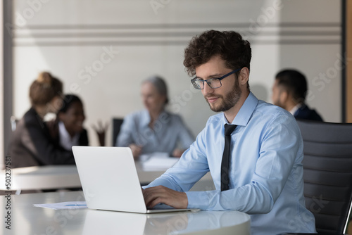 Serious young startup leader guy working on project in co-working space. Millennial businessman in glasses using laptop computer in office with corporate team in background