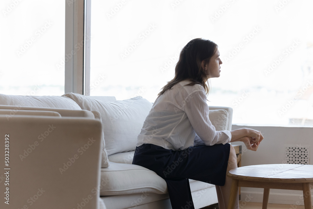 Sad depressed lonely young woman sitting on couch at home, looking at window in deep shocking thoughts, coping with loss, grief, failure, thinking over bad news, trauma, abortion, infertility Sad depressed lonely young woman sitting on couch at home, looking at window in deep shocking thoughts, coping with loss, grief, failure, thinking over bad news, trauma, abortion, infertility