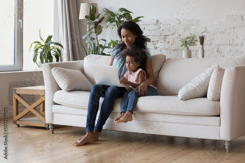 African American mom teaching little daughter girl to use learning app on laptop. Mother and kid watching cartoon movie together, enjoying leisure in living room eco interior