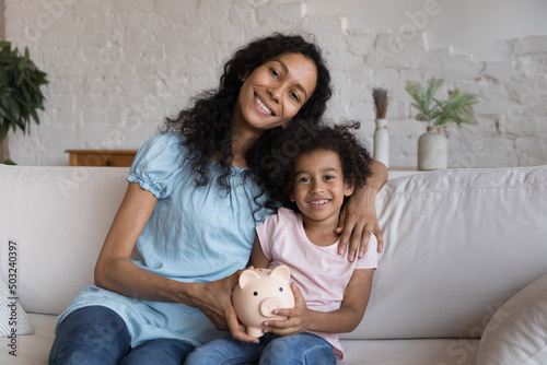 Happy mother and little daughter holding ceramic piggy bank, sitting on sofa at home, hugging, smiling, looking at camera. Family finance, reserve fund, investment, saving concept. Head shot portrait