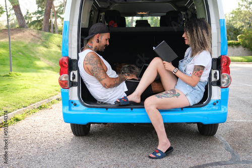 Young tattooed couple reading a book and working on a laptop sitting in the back of the van with their dogs.