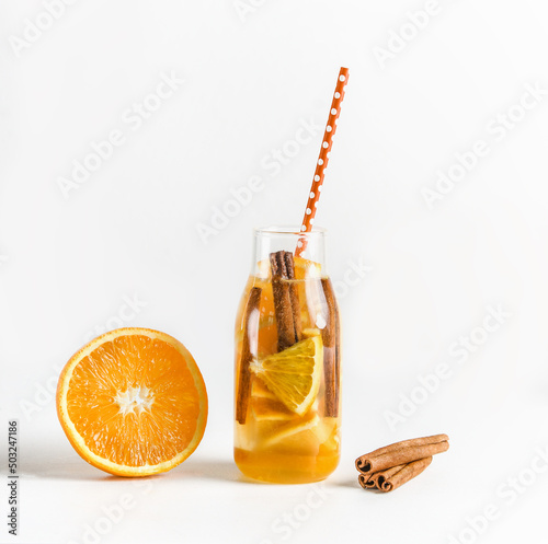 Infused water bottle with drinking straw and ingredients: orange and cinnamon sticks at white background. Summer refreshing drink with fresh fruits. Healthy lifestyle. Front view. © VICUSCHKA