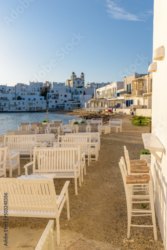 Traditional Cycladitic view with an exterior of a bar besides the sea and whitewashed houses with the christian church of kimisis Theotokou during afternoon in Naousa Paros island, Greece
