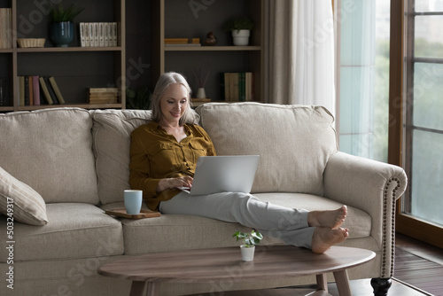 Positive mature retired grey haired woman enjoying leisure at home, resting on sofa in living room, drinking morning coffee, browsing Internet, shopping, working at laptop computer © fizkes