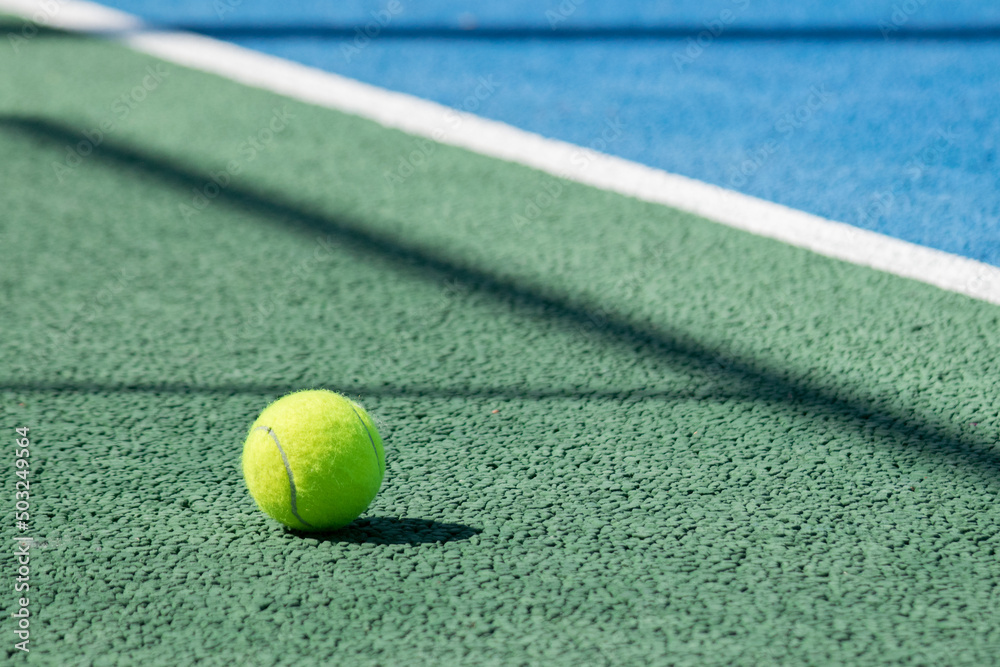 Photograph of an unmarked yellow tennis ball, on a fast tennis court of blue and green colors with black shadows and white stripes