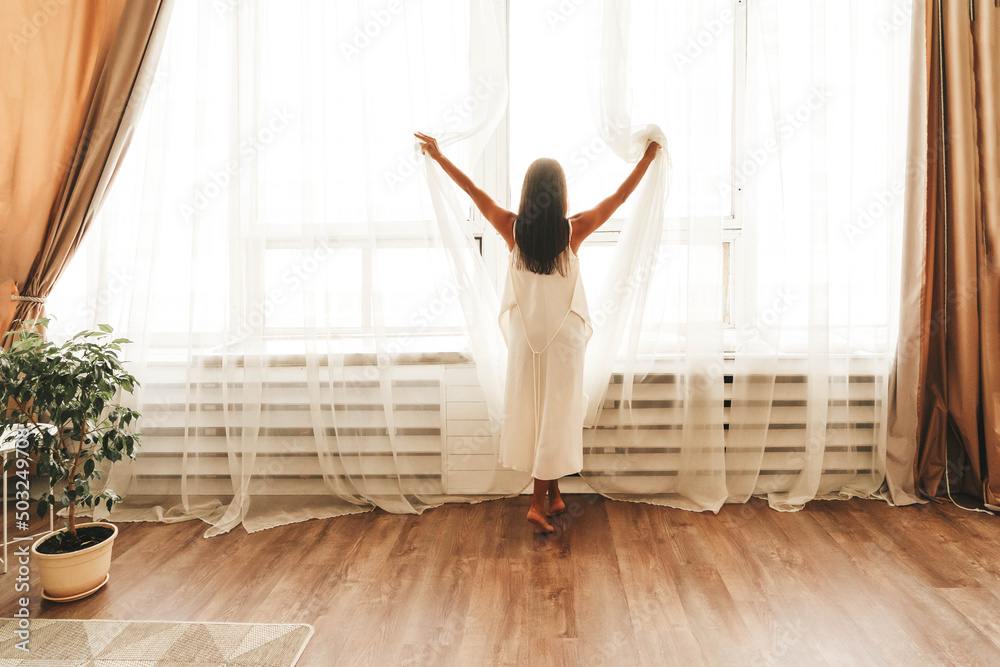 Young woman stands by large window in silk white gown after waking up at home