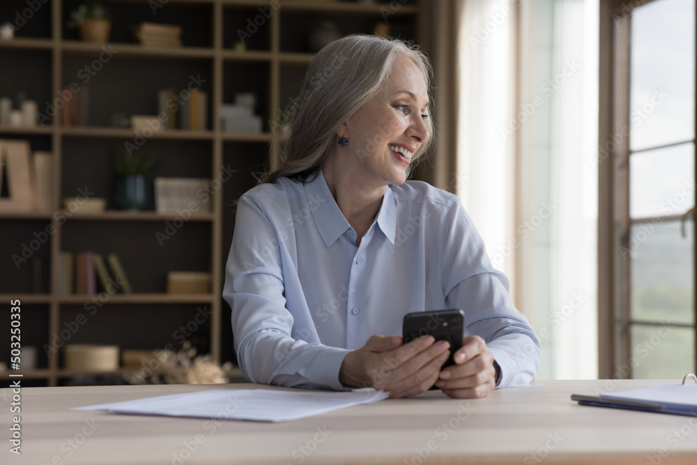Happy senior grey haired cellphone user woman holding mobile phone at home workplace table, chatting online, shopping on internet, using virtual service, looking away, smiling at good thoughts Happy senior grey haired cellphone user woman holding mobile phone at home workplace table, chatting online, shopping on internet, using virtual service, looking away, smiling at good thoughts
