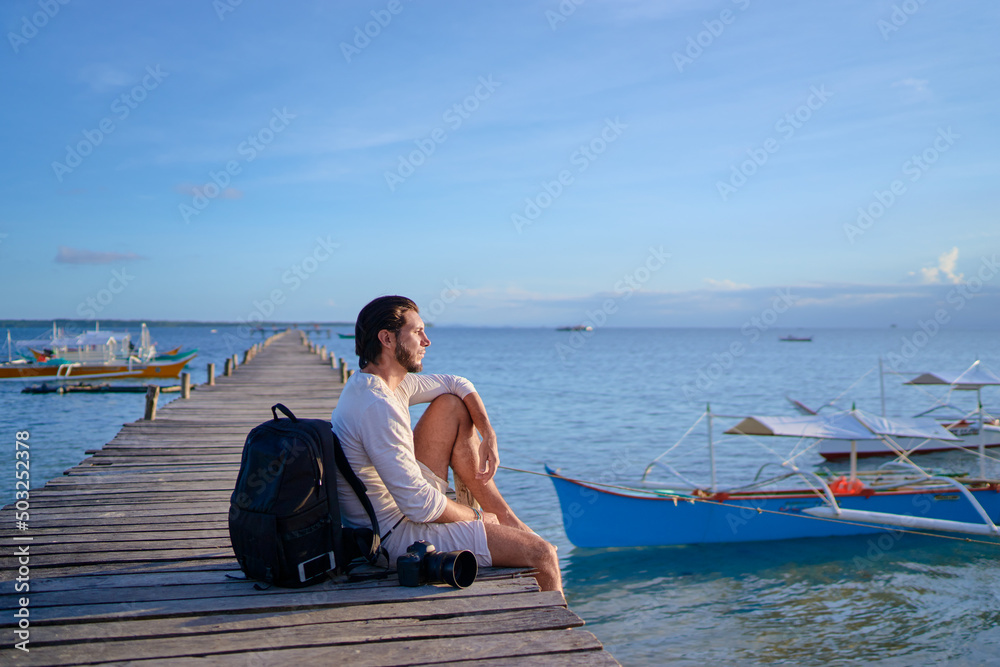 Photography and travel. Young man with rucksack holding camera sitting on wooden fishing pier with beautiful tropical sea view.