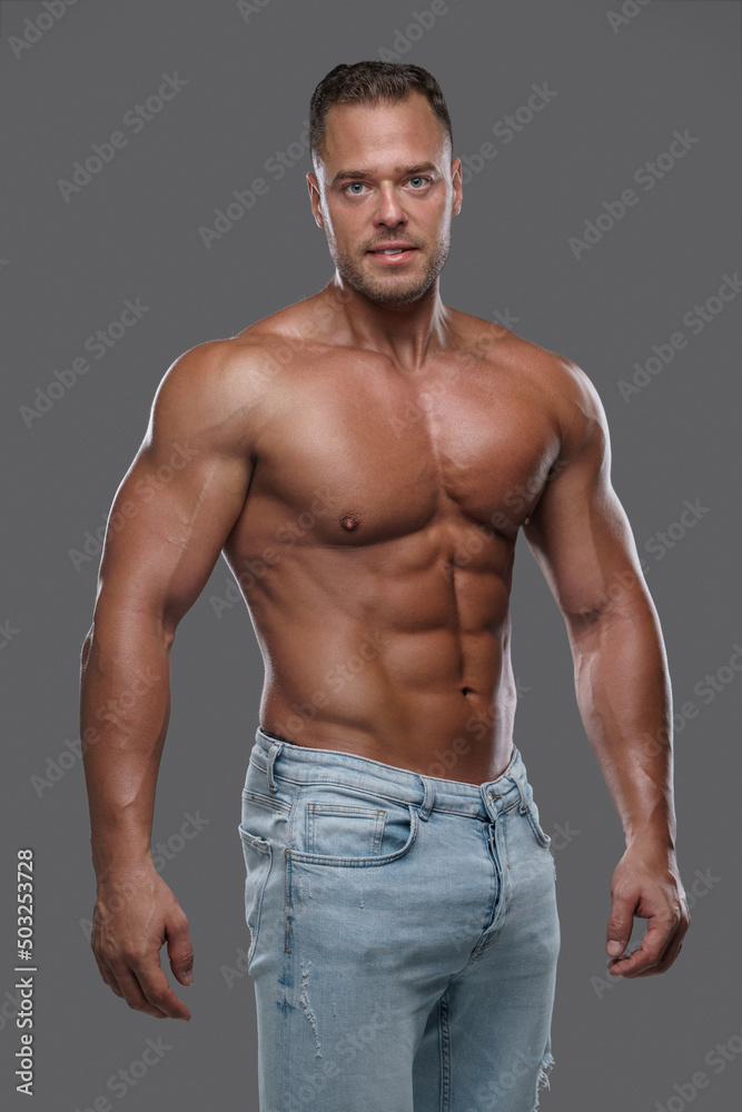 Portrait of muscular macho dressed in denim pants looking at camera against grey background.