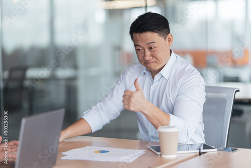 Smiling Asian businessman in meeting with colleagues in modern office showing thumbs up