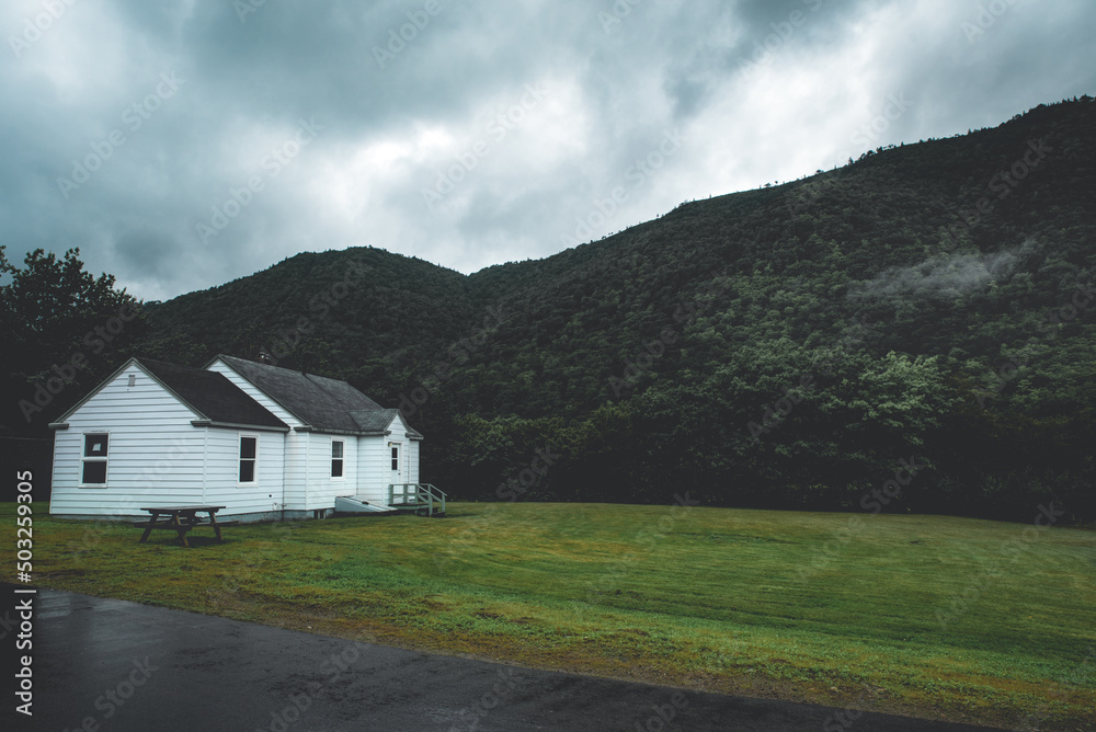 Scenic countryside landscape with green forest hills and canadian white cottage house. Cape Breton, Canada. Dark and moody filter