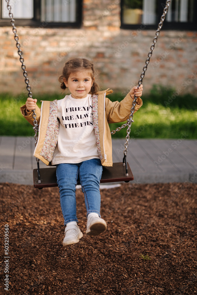 Happy little girl swing on a swing in the specially designated place in the park. Softner from sawdust underfoot Happy little girl swing on a swing in the specially designated place in the park. Softner from sawdust underfoot