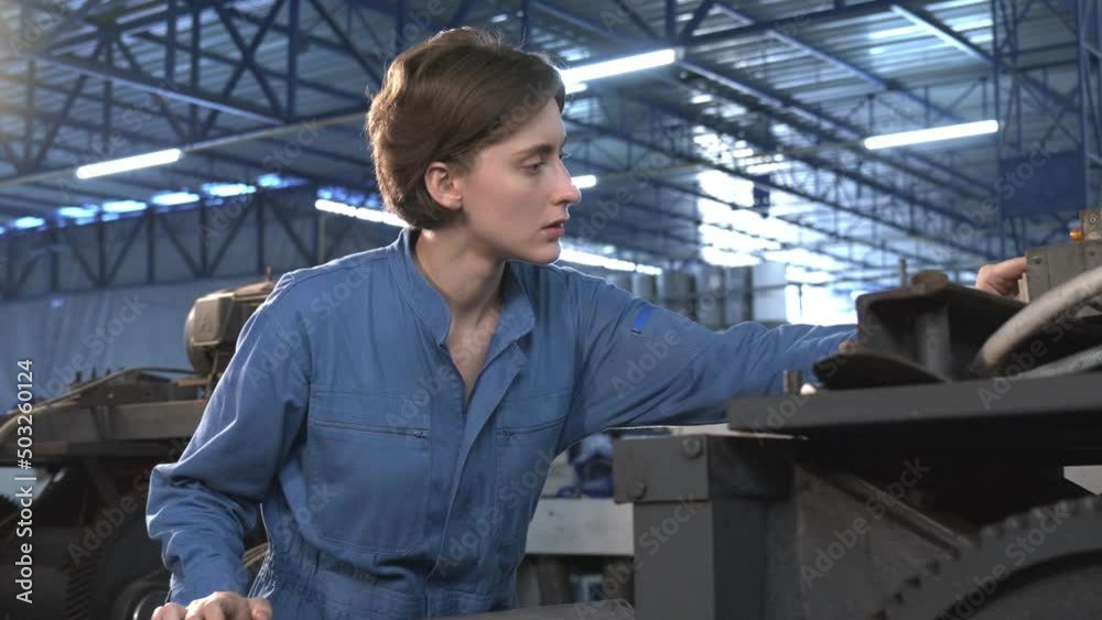 Selective focus of a beautiful short-haired Caucasian female lathe worker in a blue uniform, turning to smile at the camera while standing working with a lathe machine in a metal industry factory. Selective focus of a beautiful short-haired Caucasian female lathe worker in a blue uniform, turning to smile at the camera while standing working with a lathe machine in a metal industry factory.