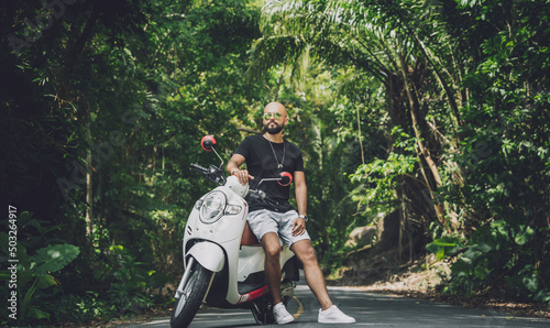 Fotografie, Obraz Stylish young man and his motorbike on the road in the jungle