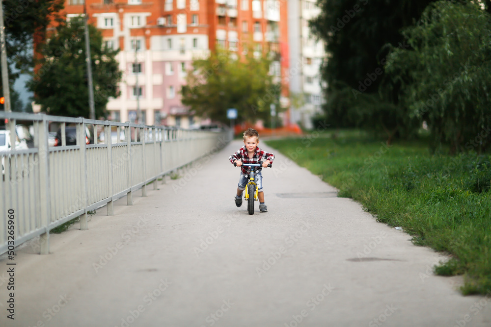 Obraz premium cute blond european boy rides balance bike on the road in city park, child rides in city in summer, lifestyle and summer vacation in town