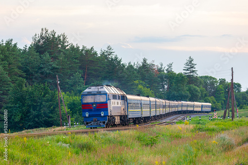 The locomotive pulls a long passenger train along the railway line through a pine forest. Summer evening lighting.