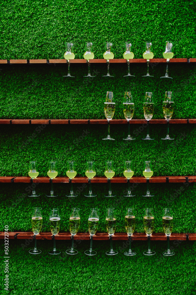 Champagne glasses and cocktails are mounted on a stand on the vertical wall of the green lawn. Decor for a wedding or party in the Italian style.