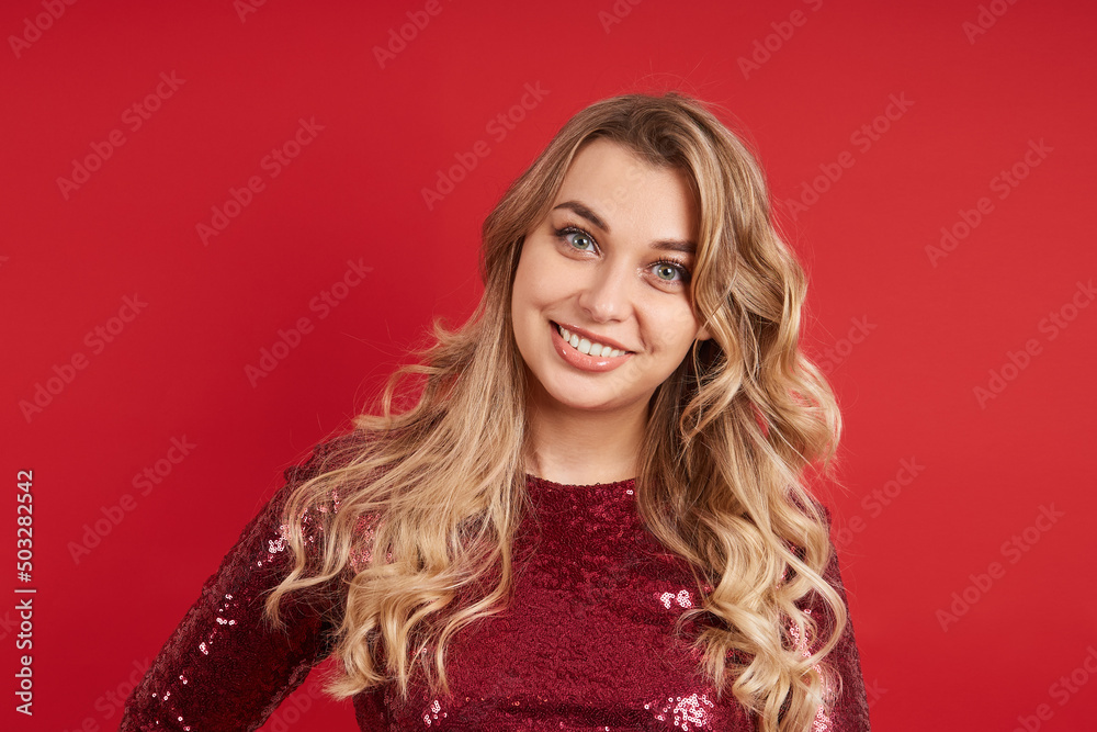 Fototapeta premium Portrait of a smiling blonde woman in a red dress on a red background looking at the camera