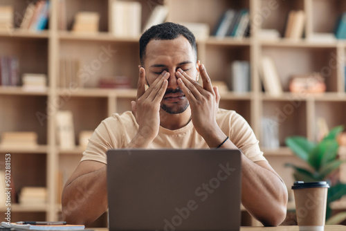 Tired arab freelancer guy sitting at workdesk in front of laptop at home and rubbing his eyes, suffering from burnout