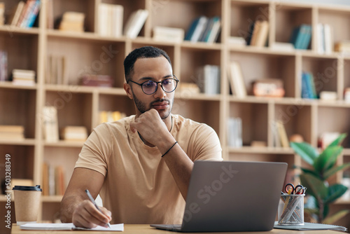 Photo Focused arab man in glasses using laptop sitting at desk and writing in notebook
