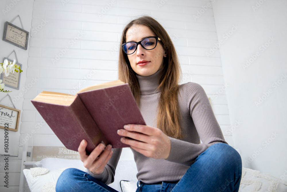 Young girl reading a book cross-legged sitting on the bed in the bedroom